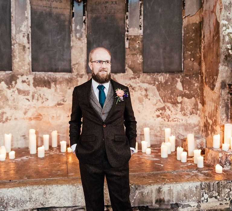 Groom in mocha brown textured suit with deep green tie and pink and purple boutonniere standing at the aisle with pillar candles 