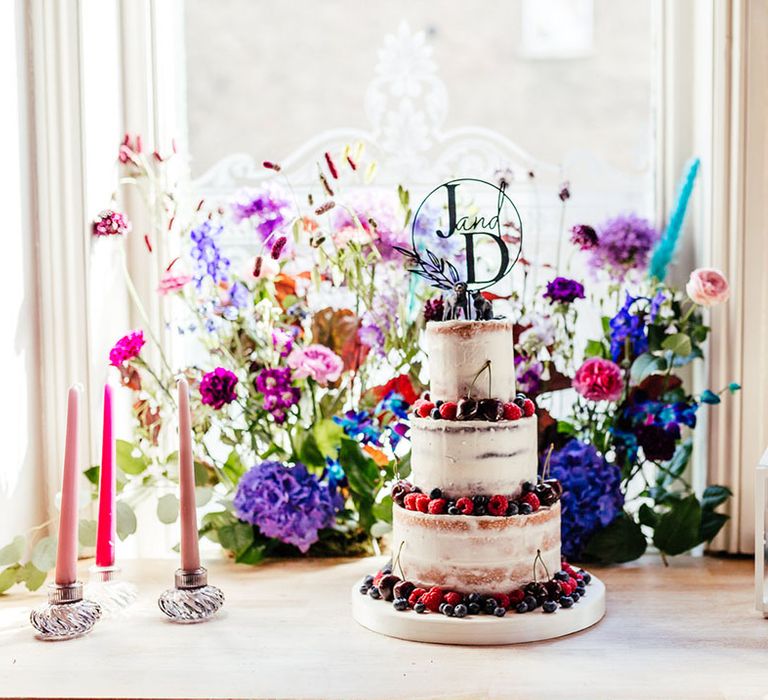 Three tiered buttercream wedding cake with laser cut personalised topper and animal wedding cake toppers with blueberry, cherry and raspberry decorations next to pink tapered candles