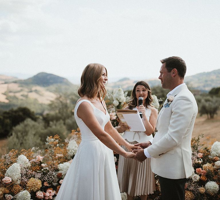 Bride in Suzanne Neville dress looks lovingly toward her husband during outdoor wedding ceremony beside wedding altar flowers