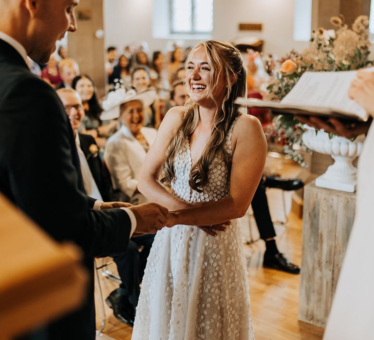 The groom in a navy suit puts on the bride's wedding ring as she laughs during the ceremony in a deep plunging neckline wedding dress 