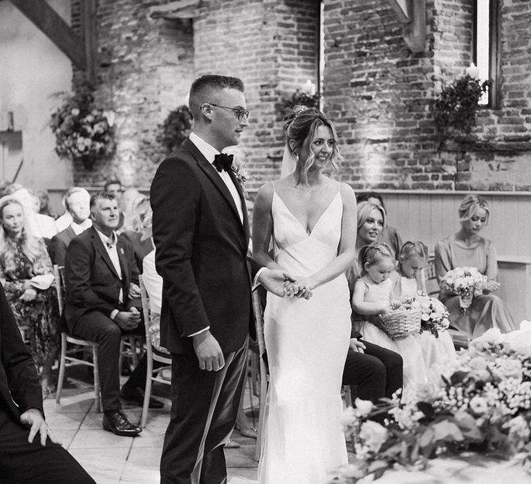 The groom in a black tuxedo stands with the bride in a satin wedding dress and v-neck style with her hair in an updo decorated with pearl hair pins