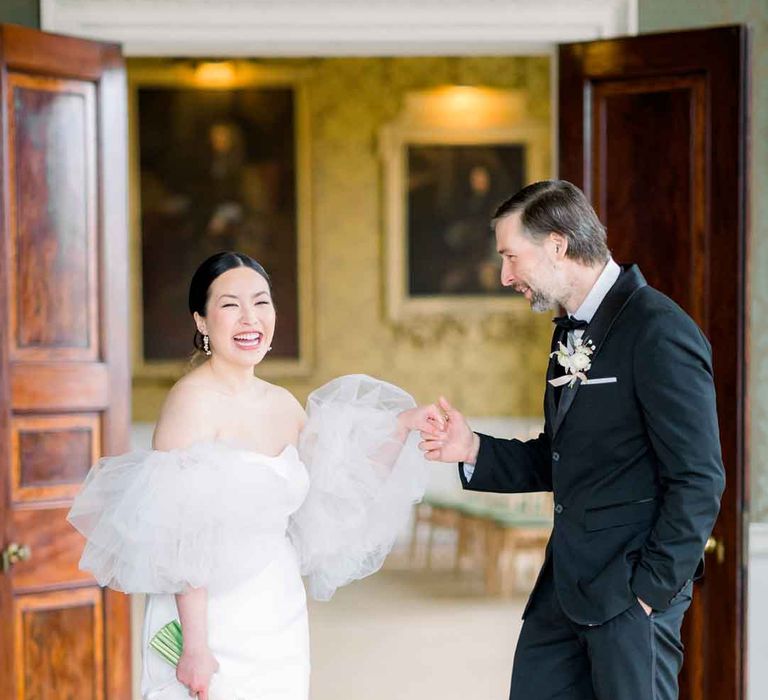 Groom in classic black tuxedo, black bowtie and boutonniere holding hands with bride in strapless wedding dress with puff tulle sleeves at St Giles House 