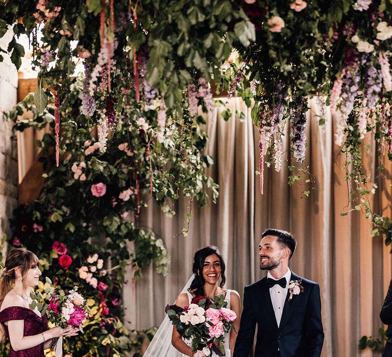 The groom in a navy tuxedo walks back down the aisle with the bride in a floral appliqué wedding dress surrounded by flowers 