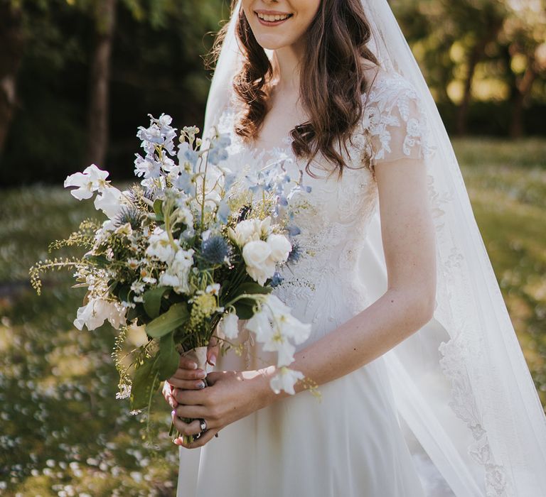 Bride in short sleeve lace wedding dress with veil holding white and blue bouquet of flowers for the wedding 