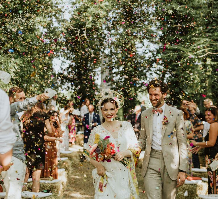 Confetti exit with bride in lace wedding dress and bridal crown carrying bright pink bridal bouquet with groom in greige suit and colourful bow tie 