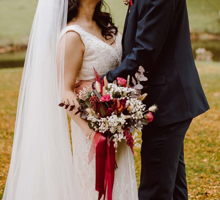 Bride in sleeveless lace wedding dress and church length red ombre veil holding white and red garden rose, peony, eucalyptus and dried foliage flower arrangements with red lace tie smiling at groom in deep blue grooms blazer, grey waistcoat, red tie, red pocket square and mixed dried flower boutonniere 