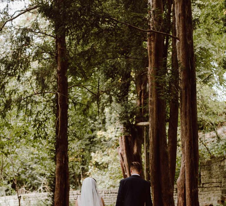 Bride in church-length red ombre wedding veil walking through woodlands of The Orangery Maidstone with groom in deep blue grooms blazer, grey waistcoat, red tie, red pocket square and mixed dried flower boutonniere 