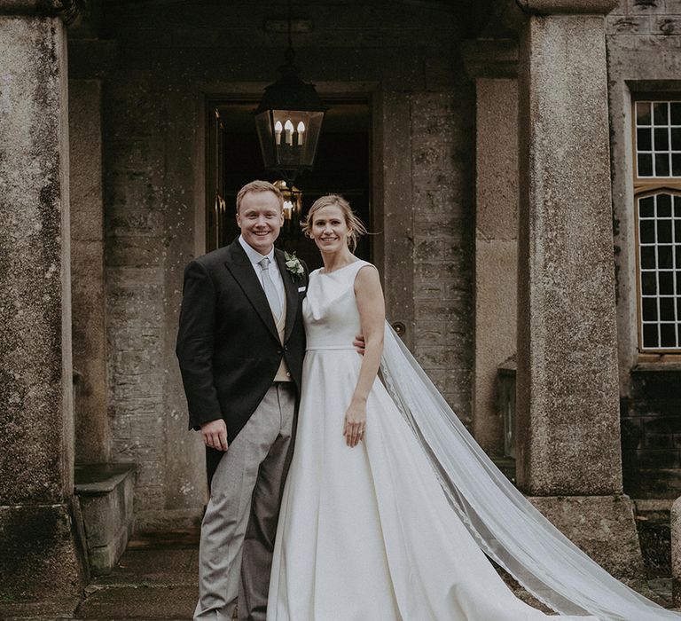 The bride wears a San Patrick wedding dress with long train and veil smiling with the groom at the entrance to the hotel venue 