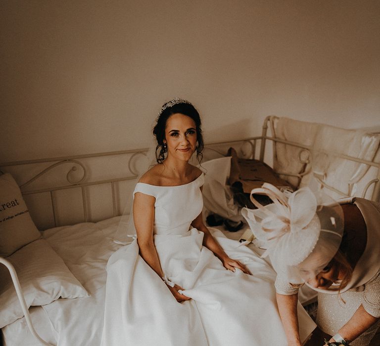 Mother of the bride helps the bride into her white block heel shoes as she sits on the bed and prepares for the wedding day 