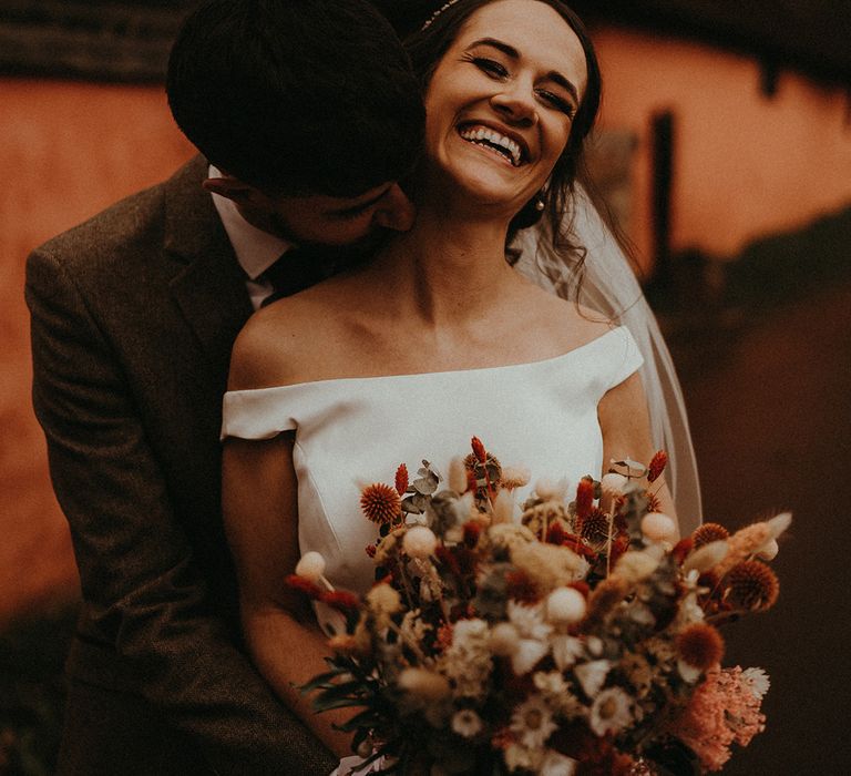 The groom kisses the bride's neck who wears an off the shoulder wedding dress holding an orange and white dried wedding bouquet 