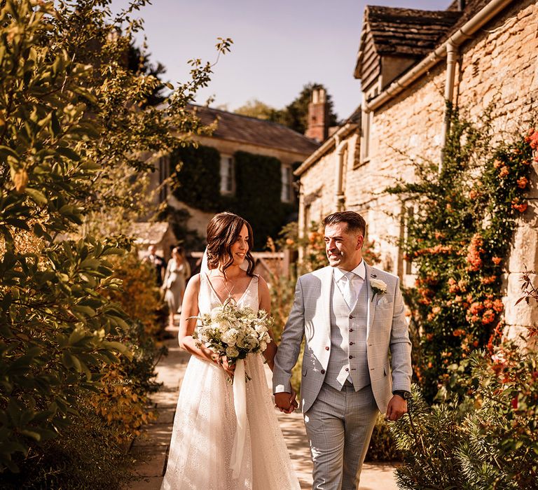 Bride in a Gabbiano sparkly wedding dress holding a white and green bouquet and her grooms hand in a light grey check suit at Caswell House 