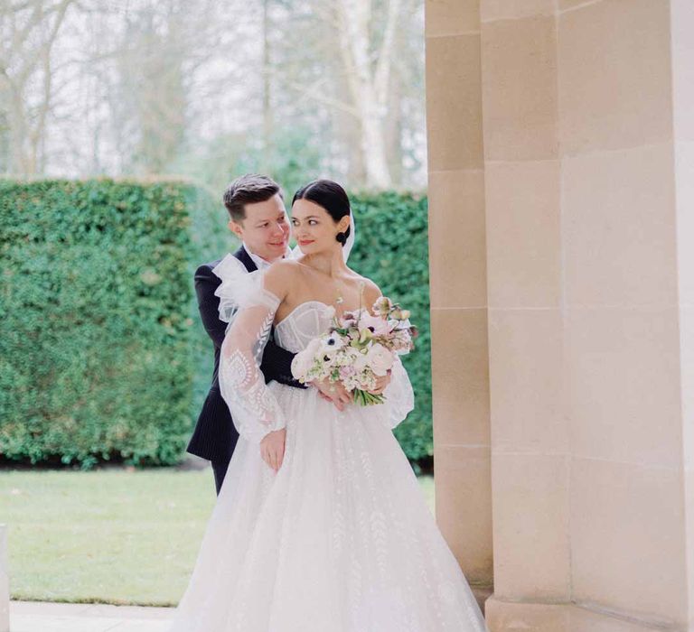 Bride in sweetheart neckline tulle mesh bridal gown with detachable puff tulle sleeves and delicate beading with cathedral length veil holding bridal bouquet with white poppies, light pink Persian buttercups, baby's-breath and foliage standing with groom in classic black grooms suit at Settrington Orangery wedding venue