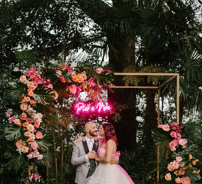 bride in a strapless wedding dress embracing her groom in a grey wedding suit in front of their gold frame altar decorated with tropical pink wedding flowers and a personalised neon pink wedding sign