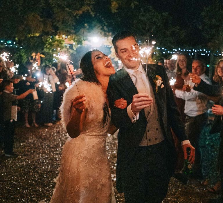 Groom in navy grooms morning suit with grey waistcoat, off-white tie and mixed wildflower boutonniere walking with bride in a-line tulle wedding dress with beaded lace applique with petals and a square neckline with side tulle cutouts and faux fur bridal coverup doing sparkler exit at South Farm wedding