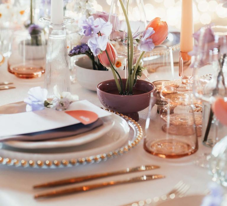 Peach and lilac wedding tablescape with spring flower centrepieces, lilac napkins, tinted glassware and gold cutlery and crockery on white tablecloth at Rackleys Barn