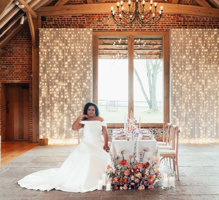 Bride in off the shoulder wedding dress with puddle train sitting at peach and lilac wedding tablescape at Rackleys Barn with wedding fairy lights wall 