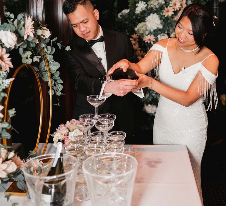 Bride in white beaded and sequined slip wedding dress standing with groom in classic black tux doing wedding champagne tower