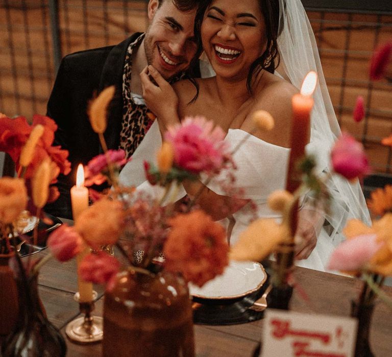 Bride in off shoulder tulle short wedding dress with church length veil sitting with groom in wet look black grooms suit with leopard print shirt and pink and orange boutonniere at rustic wedding tablescape with vinyl record place mats, gold rimmed plates, gold cutlery, colourful pink, red and orange floral centrepieces in bottles and tapered candles 