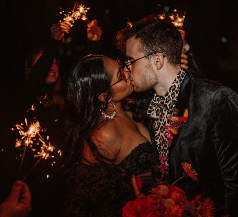 Bride in black ruffle off shoulder reception dress and red drip effect sunglasses standing with groom in black shirt and black drip effect sunglasses at German shoot with vinyl wedding arch doing fun sparkler send off