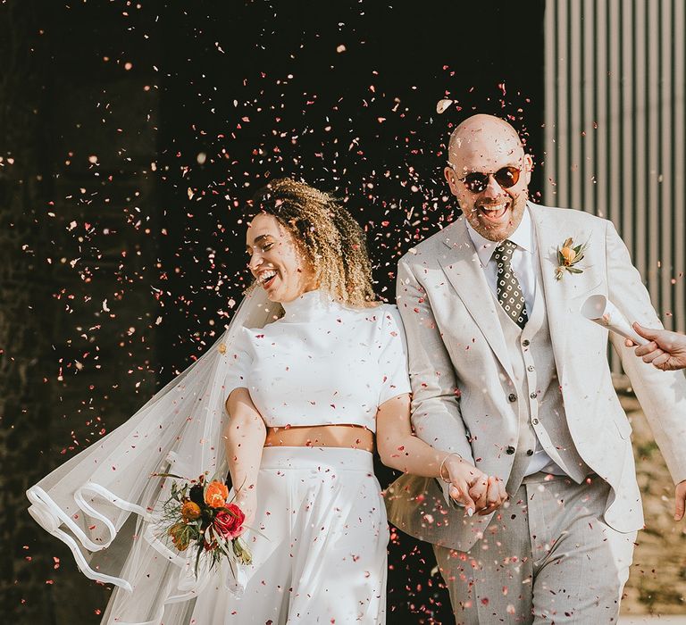 Bride in short sleeve top and trousers by Jesus Peiro with groom in cream suit walking out to confetti 