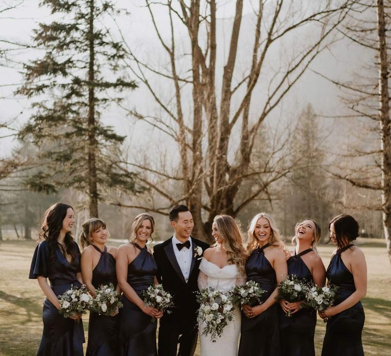 Groom in classic black tuxedo, black bowtie and white garden rose boutonniere standing with bride in off shoulder sparkly mermaid wedding dress besides bridesmaids in satin halterneck bridesmaid dresses 