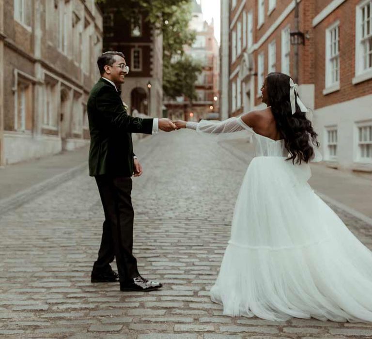 Bride in off the shoulder layered tulle wedding dress and large wedding bow hair accessory walking with groom wearing bottle green velvet grooms suit in London