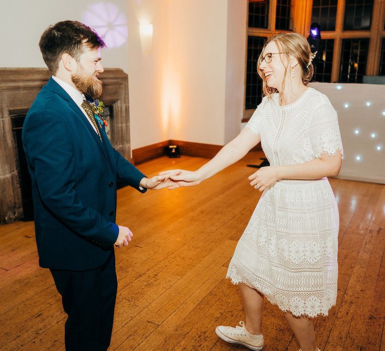 Bride in flat trainers and knee length lace reception dress dancing with the groom in his blue suit 