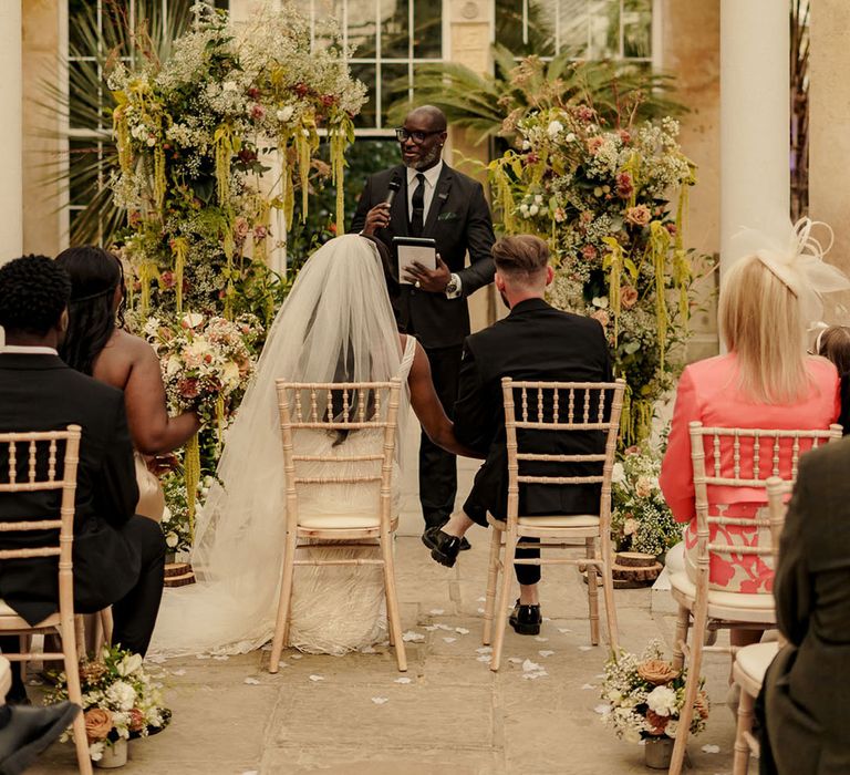 Wedding ceremony at Syon Park with the bride ad groom seated in front of floral arch decorations 