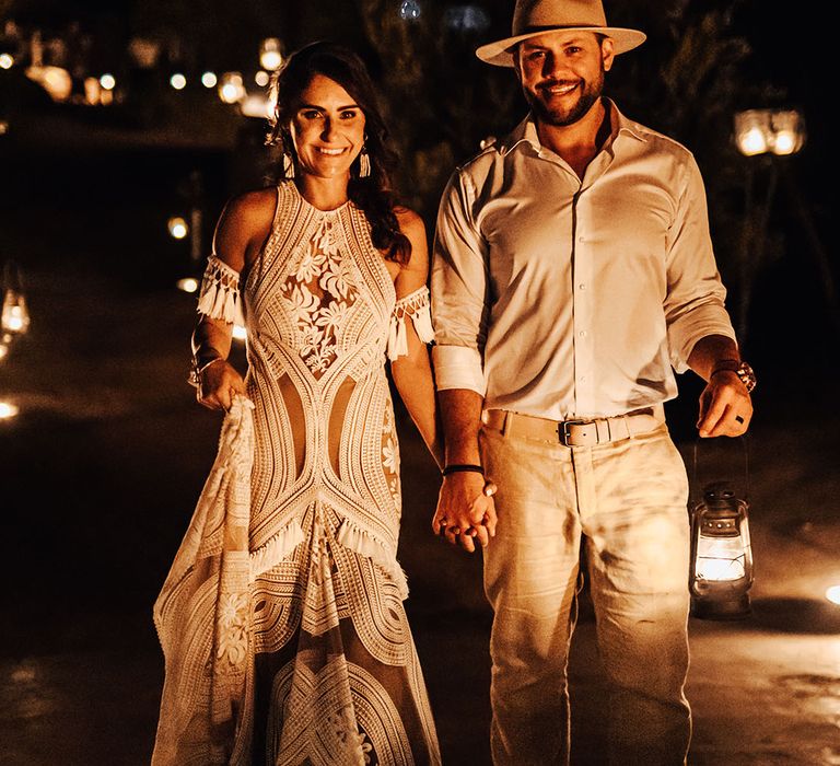 Bride in halterneck Rue De Seine boho lace wedding dress with tassels walking with the groom in beige suit and hat 