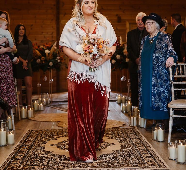 Bridesmaid in cosy burgundy velvet bridesmaid dress with cream shawl walks down the aisle at the wedding ceremony 