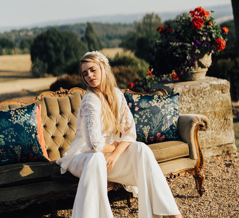 Bride in white satin jumpsuit with boho lace cover and wedges on a plush olive sofa outdoors