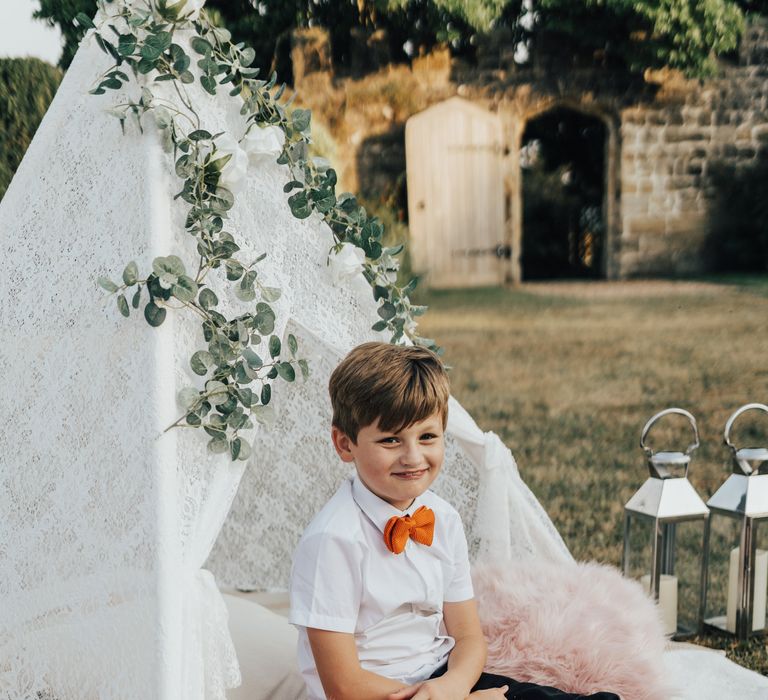 Young ring bearer inside lace teepee with orange bow tie and pink fluffy pillows and white lanterns