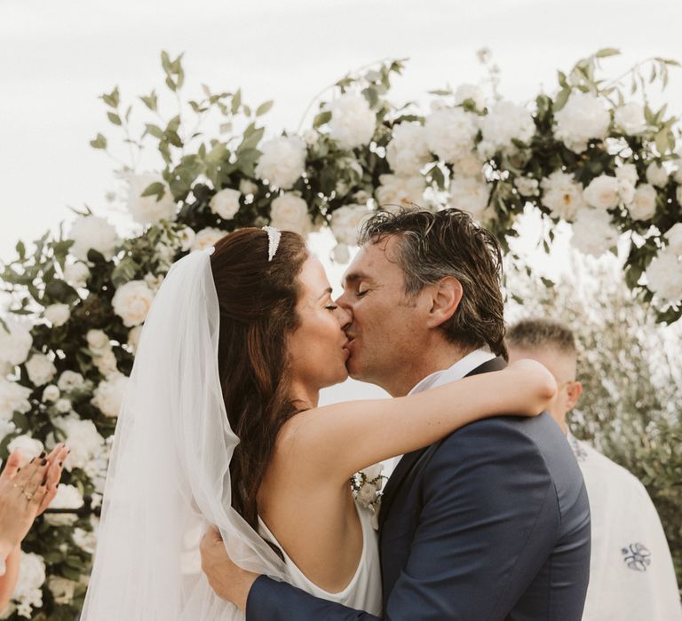 Bride and groom kissing at the altar with white flower arch 