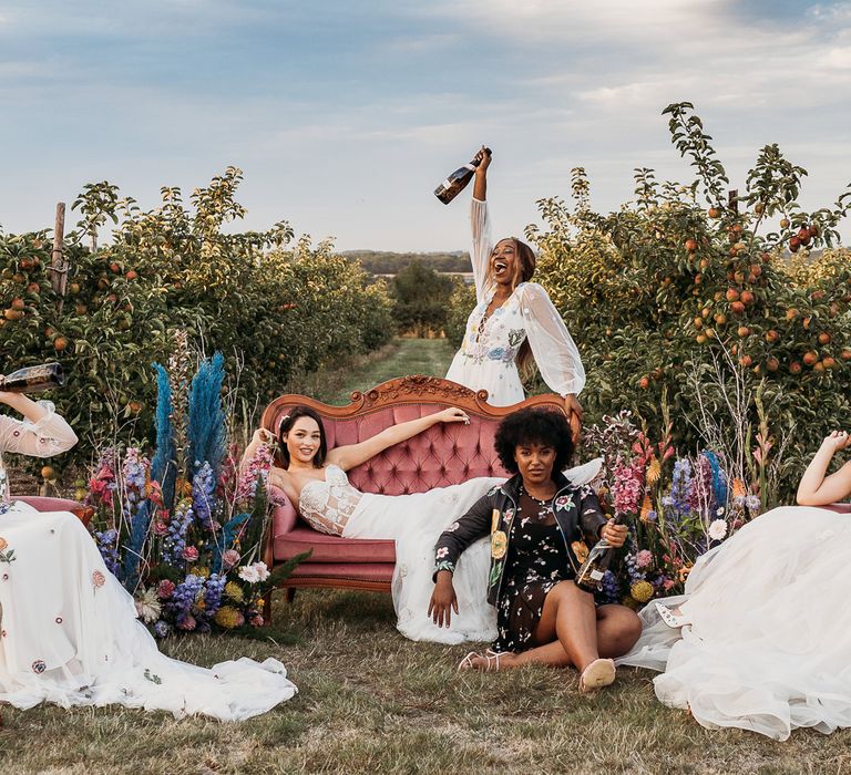 Brides in embroidered wedding dresses sitting on a pink chair surrounded by bright flowers 