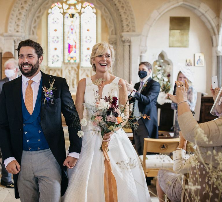 Groom in morning suit with blue waistcoat holding his brides hand in a Jesus Peiro wedding dress descending down the aisle as husband and wife