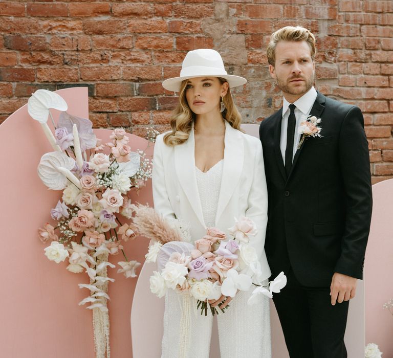 Stylish bride in a white sequin jumpsuit, blazer and bridal hat standing at the altar with her groom in a black suit