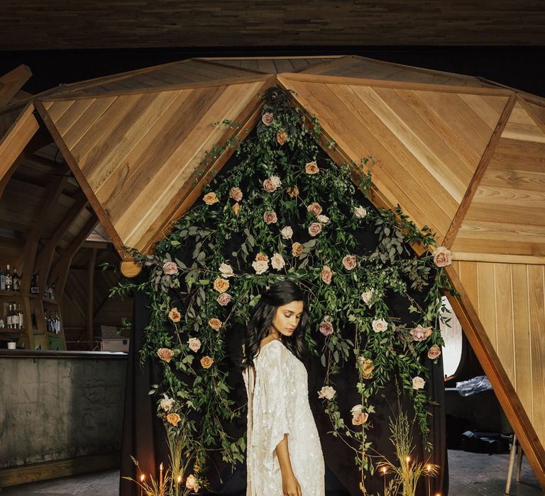 Bride in a loose fitting bohemian wedding dress with embellished detail standing at the altar with floral wall and candles 