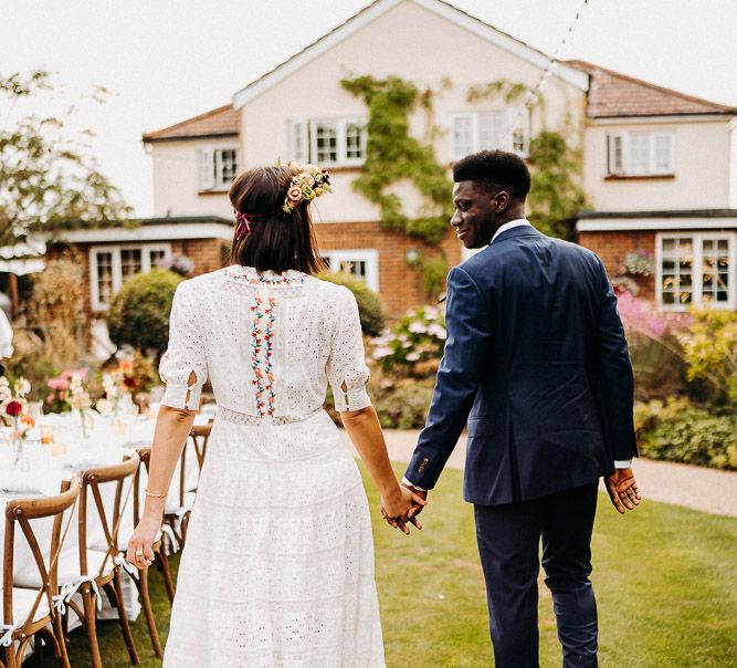 Groom in a navy blue suit holding hands with his bride in a bohemian wedding dress as they walk around the table at their outdoor reception table 