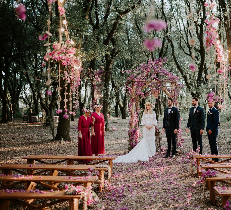 Bride in long sleeved lace boho wedding dress and flower crown stands with groom in navy suit and bow tie in front of wooden wedding arch decorated with pink flowers with bridesmaids in long fuchsia bridesmaids dresses and groomsmen in suits and bow ties