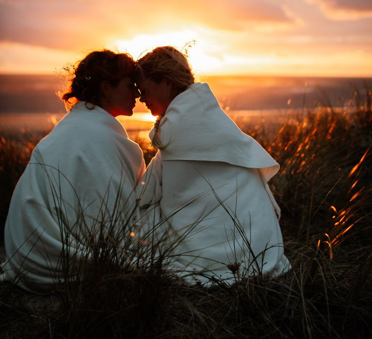 Same sex couple wrapped in blankets press foreheads together with sunset behind them