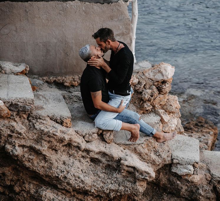 Grooms sit on the rocks to kiss in Ibiza