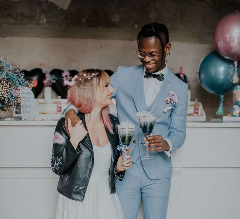 Groom in a pale blue wedding suit embracing his bride in skirt and top separates and leather jacket as they hold cocktails at the bar 