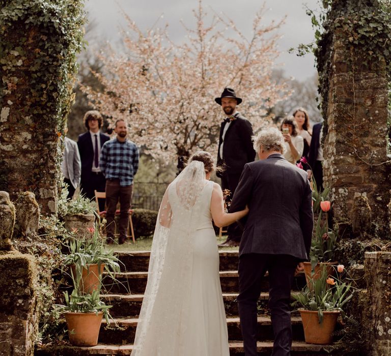 Bride in Charlie Brear wedding dress and chapel length daisy applique veil holding multicoloured bridal bouquet walks up stairs arm-in-arm with male wedding guest whilst groom in black fedora smiles