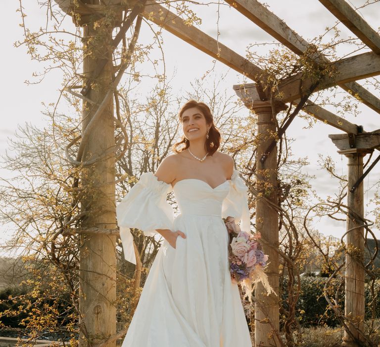 Bride stands under pergola with hands in pockets and holding pastel bouquet