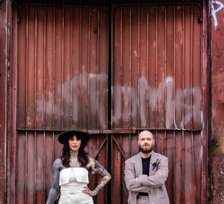 Alternative bride and groom in a strapless wedding dress and fedora hat and check suit standing neck to a corrugated iron container