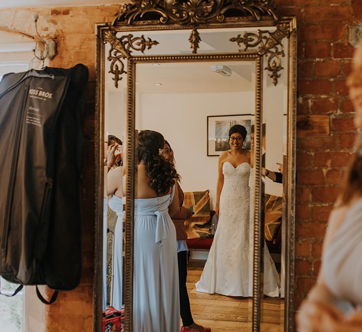 A bride in glasses looks at herself in a full length mirror in her Sophia Tolli gown.