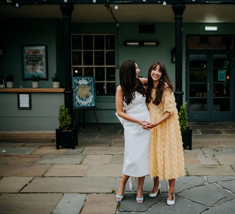 Bride in white strapless Rebecca Vallance dress and blue slingback heels stands with arm around wedding guest in midi yellow short sleeved dress with feather detailing in courtyard at Hotel du Vin wedding reception in Harrogate