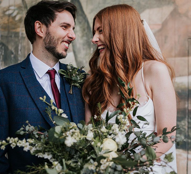 Groom in a navy suit with red satin tie laughing and smiling with his bride in a satin slip Grace Loves Lace wedding dress on stage at The Larmer Tree's outdoor theatre 