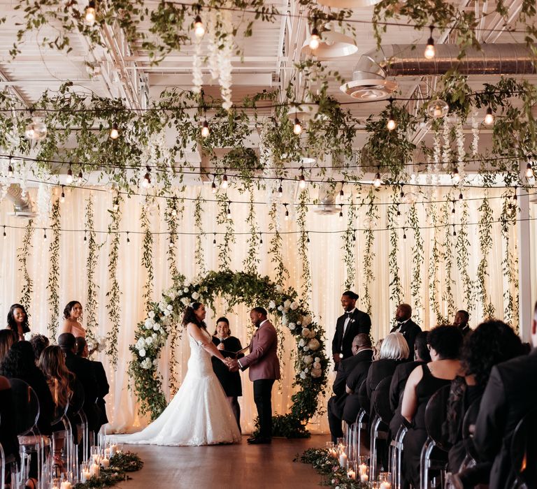 Bride & groom during wedding ceremony in front of floral archway and curtain whilst guests are seated on clear chairs and candles line the aisle