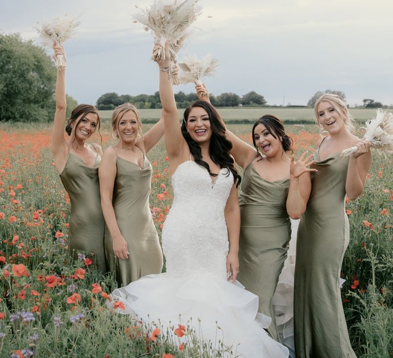 Bride in a sweetheart neckline fitted wedding dress with fishtail skirt holding her neutral dried flower bouquet in the air flanked by her bridesmaids in satin green dresses 